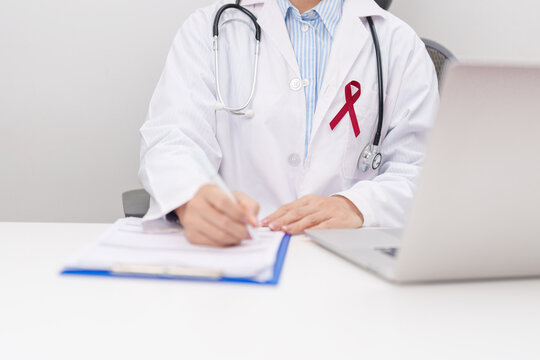 doctor using laptop and report notes on notebook. burgundy ribbon concept of March Multiple Myeloma Cancer Awareness month, September Sickle Cell Anemia, Headache and Migraine week, April Meningitis