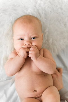 Baby 5-6 months old lies on back on a soft white blanket and sucks fingers in a calm home scene