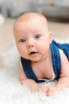 Baby 5-6 months old in blue overalls lies on tummy on a white blanket at home and looks at camera