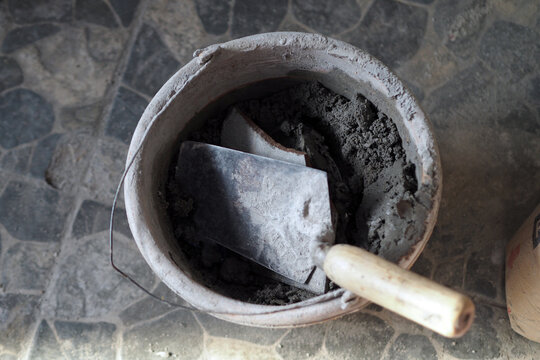 top-down view of a bucket filled with dry cement powder, debris, and a construction trowel resting inside.