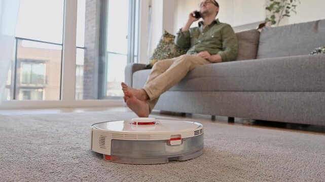 Smart vacuum cleaner cleans the carpet while man sits on the sofa. A smart home, modern technologies,housekeeping concept