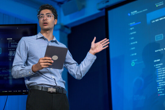 A male analyst pointing at a specific metric on his monitor points precisely at the figure that has triggered the alert at a developer desk washed in blue light from surrounding displays