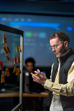 A male scrum master reviewing a wall of task cards with his team holds a tablet in one hand while rearranging the sticky notes in a quiet corporate office in a modern technology company