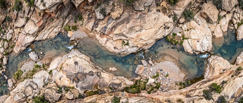Aerial view of a winding river flowing through a rocky canyon, featuring tan sandstone cliffs, clear blue water, and small patches of green vegetation along the riverbanks