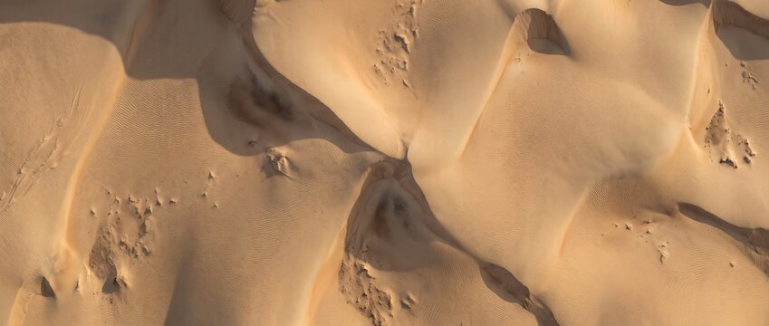 Aerial view of golden sand dunes in a vast desert landscape, featuring soft curves, sharp ridges, and deep shadows cast by the sun across the undulating terrain
