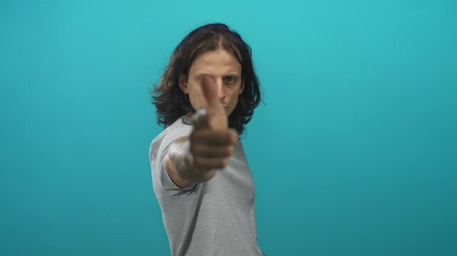Man with long hair pointing index finger at camera with outstretched hand in teal studio; confidence focus.