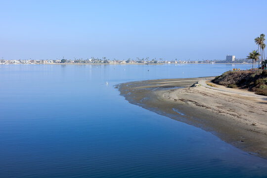 Tranquil blue Mission Bay flows gently beside a curved sandy beach with distant coastal buildings and palm trees under clear skies, California, San Diego
