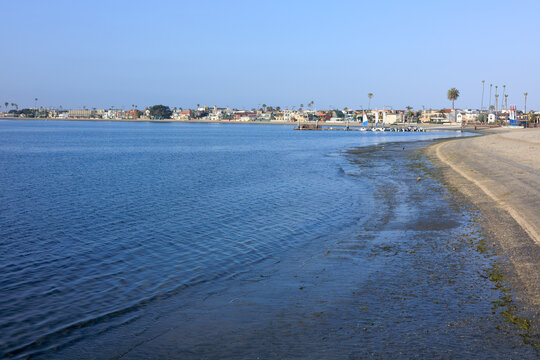 A calm blue Sail Bay water curves along a sandy shoreline with coastal houses, a distant pier, and palm trees under clear sky, San Diego, California