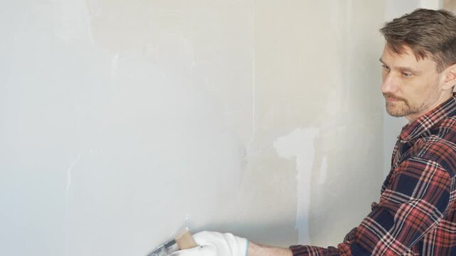 Male construction worker spreading plaster on white wall, wearing protective gloves during home renovation project with putty knife