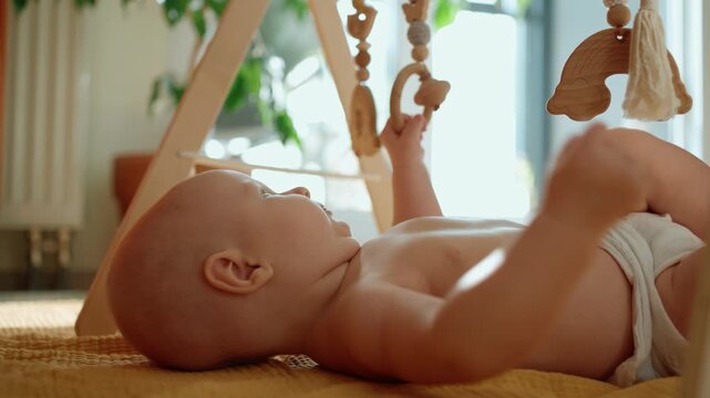 Adorable baby boy lying on a floor mat and playing with a wooden play gym at home, his tiny hands reaching for the hanging toys, promoting early sensory development and learning