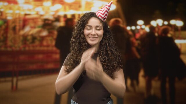 Woman wearing a chevron party hat covers mouth with hands while smiling at a carnival carousel at night; celebration joy.