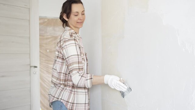 Woman applying plaster to wall with putty knife, wearing protective gloves and creating smooth surface during home renovation work
