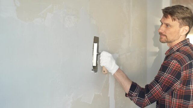 Male construction worker wearing protective gloves, spreading plaster evenly on wall surface using putty knife, renovating interior with professional precision and smooth technique