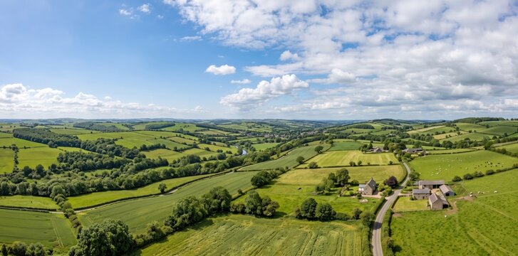 Panoramic aerial view of the lush green rolling hills of the devon countryside, featuring patchwork agricultural fields, hedgerows, scattered farmhouses, and a winding road under a bright blue