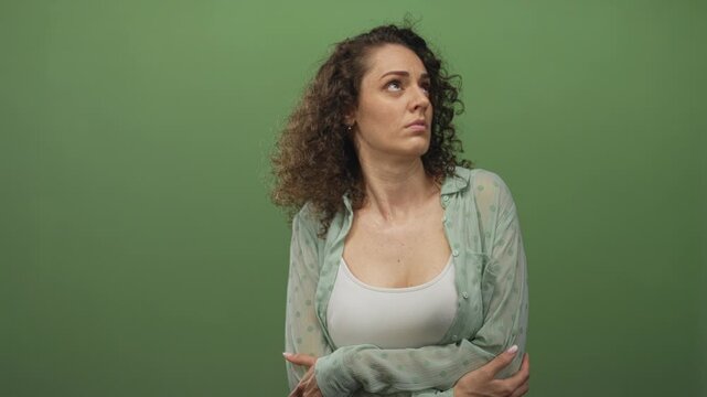 Woman with arms crossed exposing chest and shoulders in green studio, wearing white tank top and sheer button shirt, looking to the side while holding herself; vulnerability introspection concern.