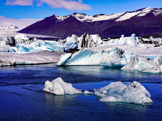 Sculpted blue and white icebergs drift across the waters of Jökulsárlón glacier lagoon against a backdrop of snow-streaked volcanic mountains in southeastern Iceland © Gregory
