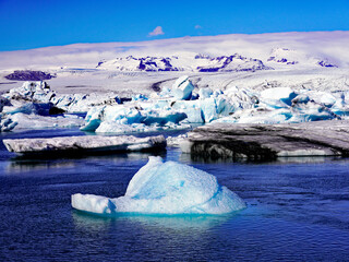 A floating blue iceberg drifts in the foreground across Jökulsárlón glacial lagoon with vast ice floes and the snow-covered Vatnajökull ice cap in the distance in southeastern Iceland © Gregory