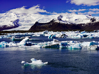Numerous icebergs drift across the waters of Jökulsárlón glacial lagoon in front of massive snow-covered peaks and the Vatnajökull ice cap under a blue sky in southeastern Iceland © Gregory