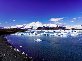 Bright blue and white icebergs drift across Jökulsárlón glacial lagoon beneath massive snow-covered glacier peaks and a deep blue sky in southeastern Iceland © Gregory