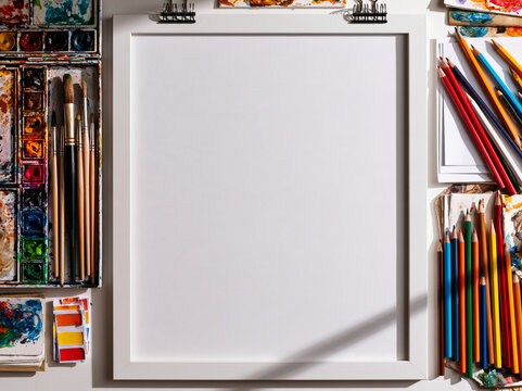 An overhead shot of art supplies laid out on a flat surface, featuring a blank canvas clipboard, watercolors, brushes, and colored pencils