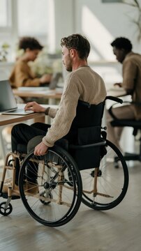 A person in a wheelchair works at a laptop in an open bright office space with clean modern furniture and soft daylight coming through the wide windows behind the desk