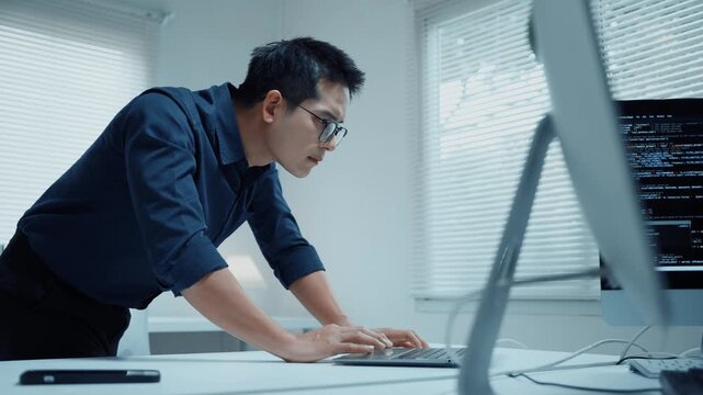 Focused asian programmer developing software and typing code on laptop standing at his desk in a modern office