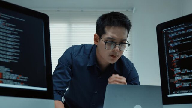 Focused asian programmer writing code and debugging software on multiple computer screens in a modern office environment