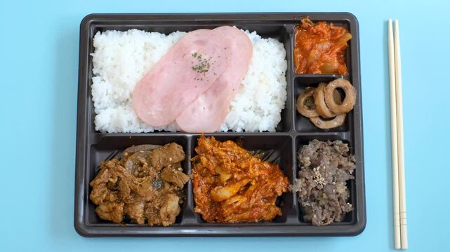 Korean lunch box with rice, meat, and kimchi in plastic packaging on a blue background close-up, top view.