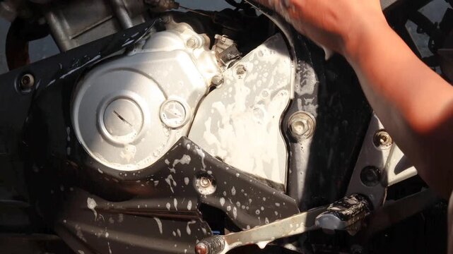 Close-up of a person's hand cleaning a motorcycle engine and body with a soapy sponge during vehicle maintenance and washing at home.
