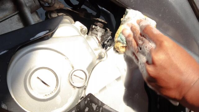 Close-up of a person's hand cleaning a motorcycle engine and body with a soapy sponge during vehicle maintenance and washing at home.