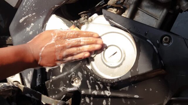 Close-up of a person's hand cleaning a motorcycle engine and body with a soapy sponge during vehicle maintenance and washing at home.