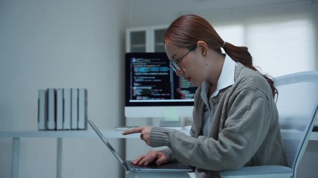 Asian female programmer developing software and debugging code on laptop and computer screen working in a modern office at night