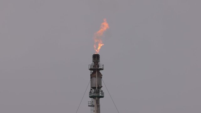 Flaming oil refinery flare stack with fire burning gases against a grey overcast sky. Industrial metalwork, moody lighting, and static wide angle capture energy industry process.