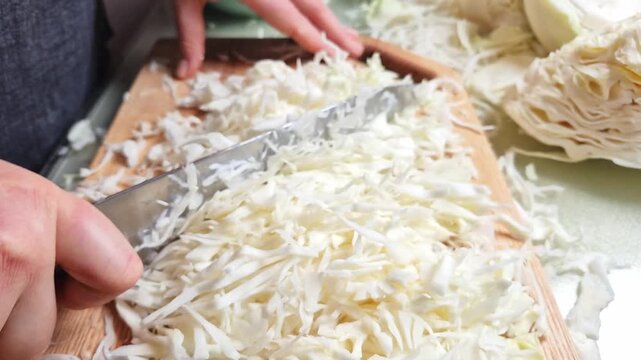 A person is meticulously slicing a fresh head of cabbage into very fine shreds using a sharp knife on a wooden cutting board, captured in slow motion