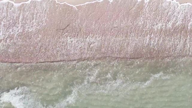 Aerial view of ocean waves washing over a sandy beach, creating beautiful natural patterns of white foam and seawater along the shoreline.