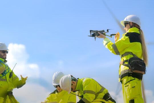 Bearded technician with a blue rope coil lifts a pointing finger toward the drone that a blonde colleague balances on her fingertips above two crouching crew members