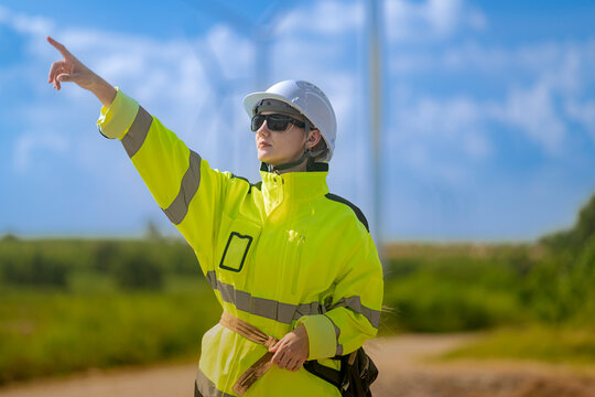 Female wind technician points up and to the left with her mouth slightly open as if calling out, a green hedge and warm summer field softened behind her, a breezy day of fieldwork