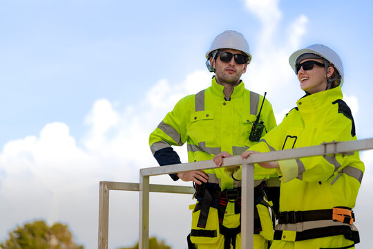 Male and female technicians share a laugh at the rail of a service platform, sunglasses on and white hard hats bright against the pale cloud with clean power turning behind