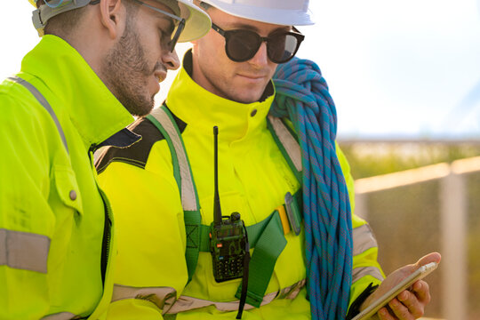 Very close portrait of two technicians, a black two-way radio clipped to the harness of the man in front while a colleague leans in behind his right shoulder in bright sun