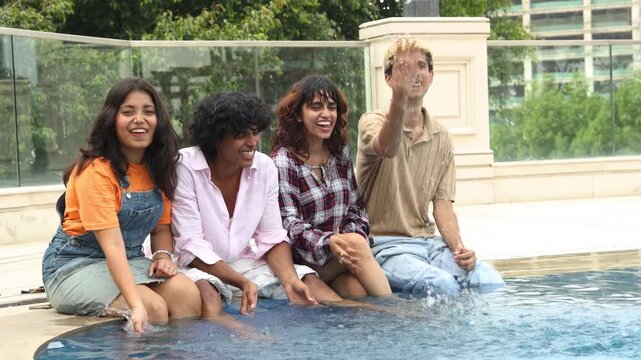 Diverse young indian asian Gen Z friends sitting together with their legs in swimming pool enjoying happy summer day outdoors splashing water . Travel and Holidays..Youth Culture.