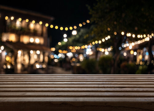 Empty wooden table sits before blurred night market lights. Table surface provides open copy space, and warm bokeh strings lead past out of focus stalls, giving peaceful evening ambience.