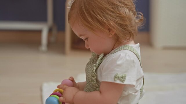 Toddler girl holds colorful ring rattle in hand while sitting on play mat in nursery; curiosity innocence play.