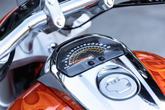 Motorcycle instrument cluster and fuel tank gleam in close up. Chrome handlebars frame speedometer on orange bodywork, showing sleek mechanical detail and polished design with focused calm.