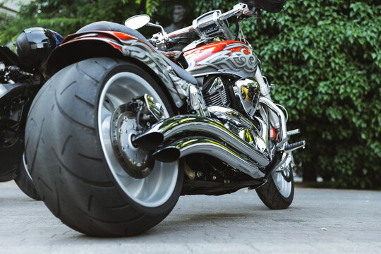 Custom cruiser motorcycle rests on paved driveway, showing oversized rear tire, chrome exhaust pipes, and red silver flame paint. Static bike sits against leafy green hedge in gleaming display.