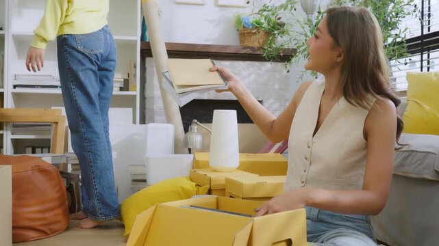 Happy two Asian women friend picking up things out to a paper box while preparing to move in of the new home. Relocation concept