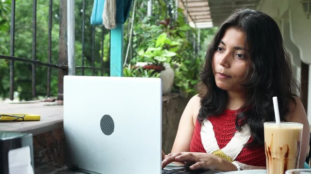 Young indian gen z woman working on laptop while enjoying a snack and a coffee milkshake at a restaurant or cafe, Concept of modern digital nomad lifestyle and remote work flexibility, freelancing