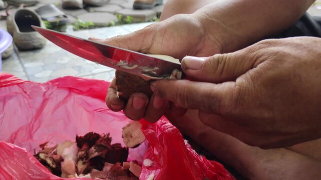 A close-up shot of a pair of hands peeling raw cassava tubers with a knife over a red plastic bag filled with pieces of cassava, under natural outdoor light.