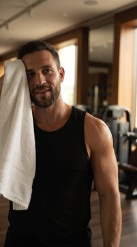 Portrait of smiling muscular man wiping sweat from face with white towel after gym workout, black tank top, rest and recovery concept.