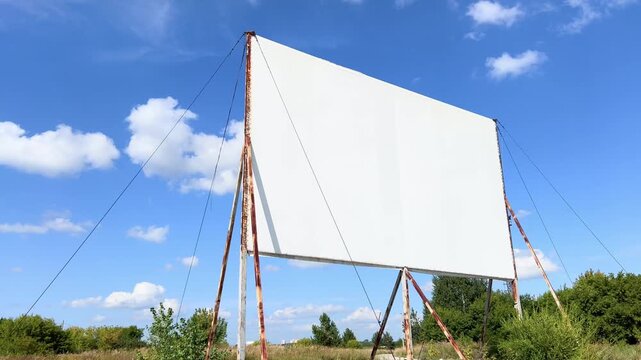 Large white outdoor projection screen billows in wind above a green field under blue sky