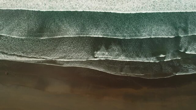 Aerial top view of ocean waves forming patterns on dark sandy shore